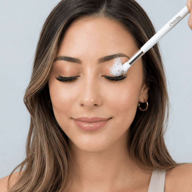 women softly smiling, washing her eyelid with a white foam and a prolong lash eyelash extension cleansing brush on a blue background