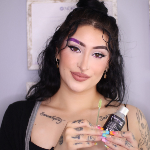 Woman holding a bottle of prolong lash product with visible tattoos and colorful makeup.