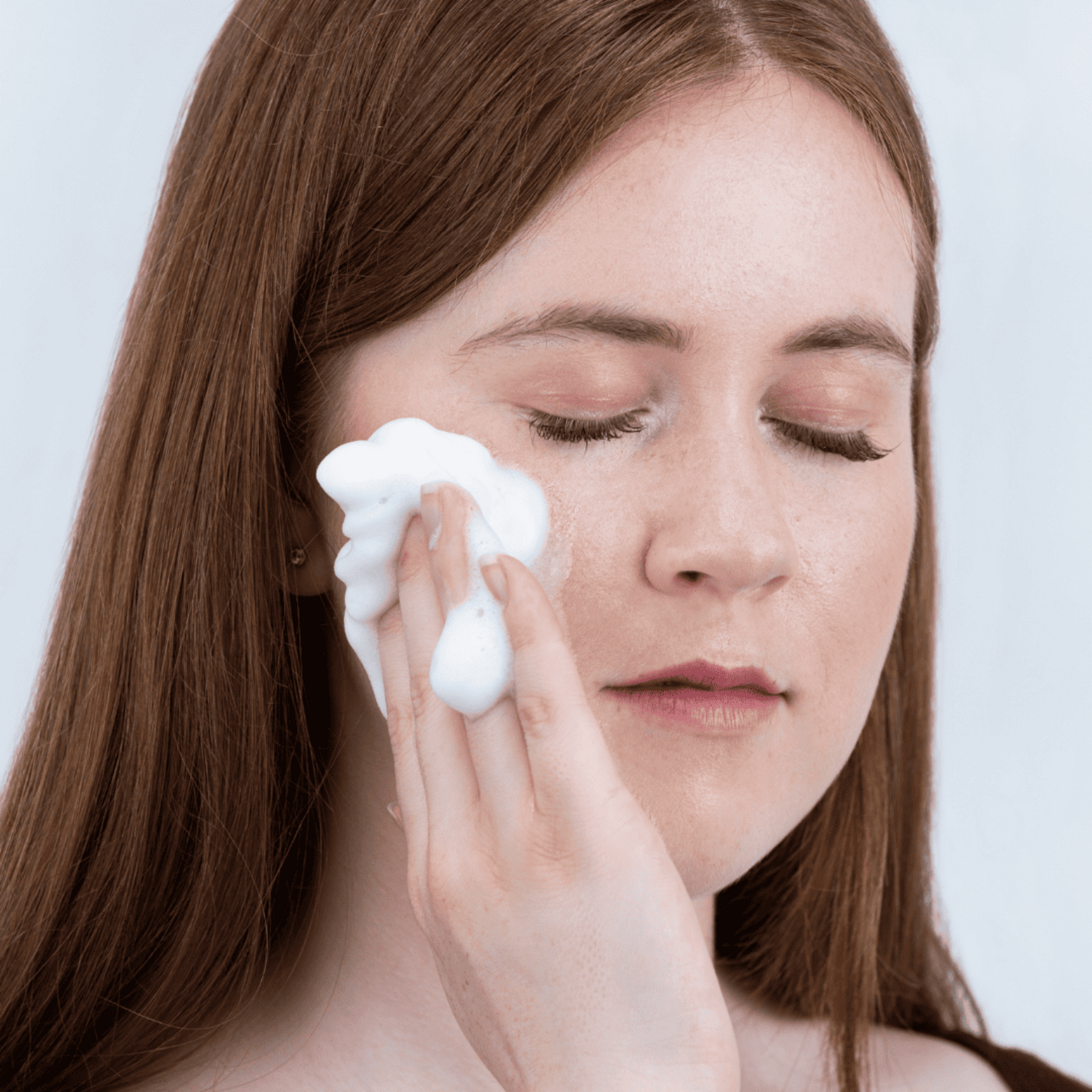 Woman applying foam cleanser to her face against a plain background