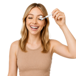 A woman with blonde hair smiling while using a white eyelash cleansing brush on her eye, wearing a beige ribbed tank top against a solid white background.