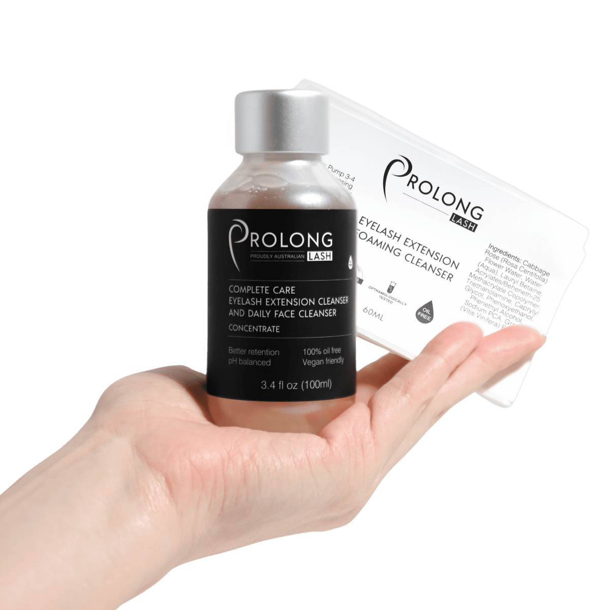 A hand holds a small bottle of Prolong Lash Complete Care Eyelash Extension Cleanser and Daily Face Cleanser Concentrate next to a prolong lash retail label on a solid white background.