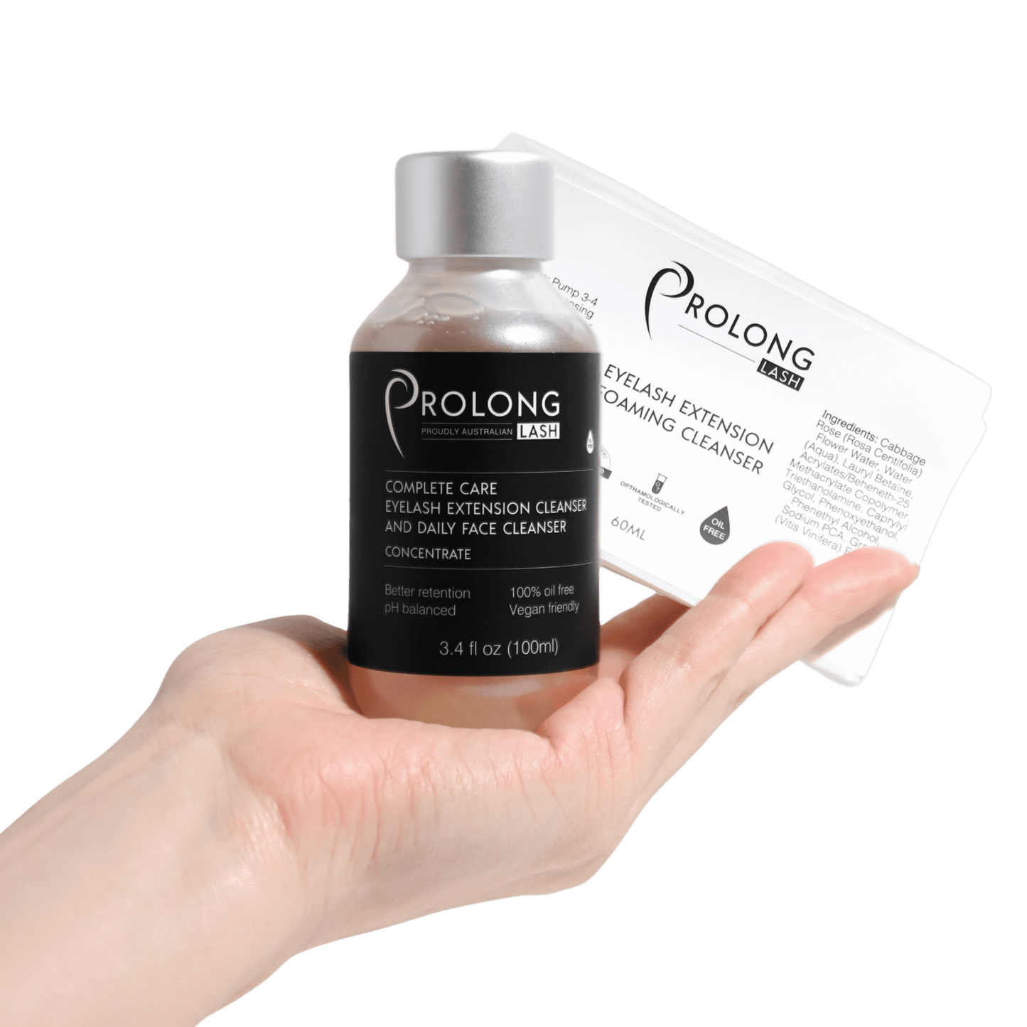 A hand holds a small bottle of Prolong Lash Complete Care Eyelash Extension Cleanser and Daily Face Cleanser Concentrate next to a prolong lash retail label on a solid white background.