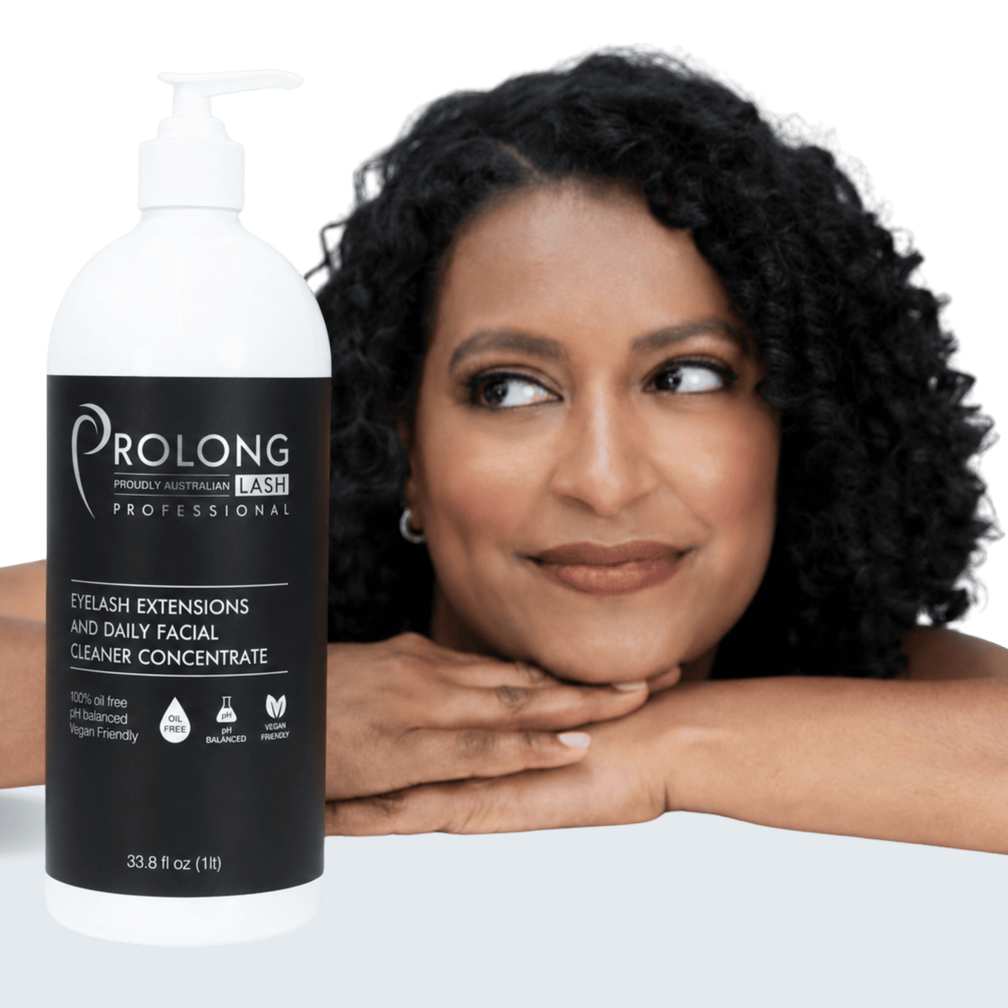 A bottle of Prolong Professional eyelash extensions and daily facial cleaner concentrate shown in front of a smiling woman with dark curly hair resting her head on her hands against a clean white background.