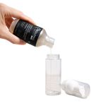 A hand pours prolong lash concentrate from a black-labeled bottle into a clear cylindrical pump bottle, with the white pump cap placed beside it on a white background.